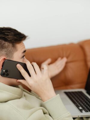 A young man in a hoodie talks on his phone while using a laptop on a brown couch.