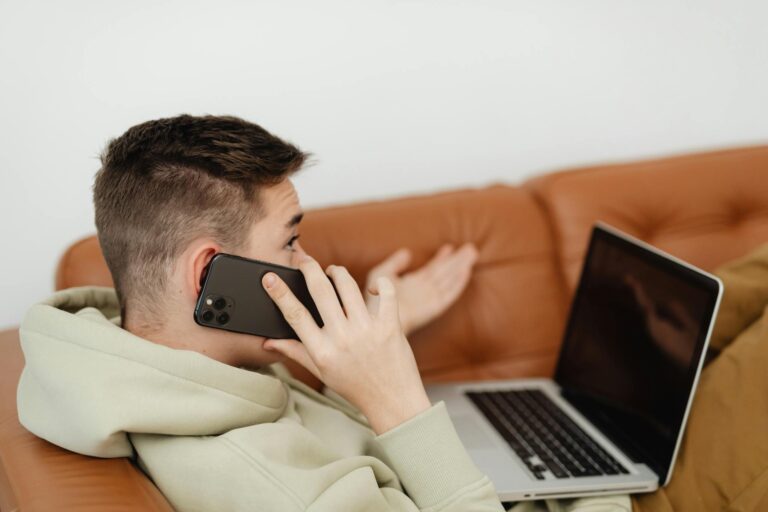A young man in a hoodie talks on his phone while using a laptop on a brown couch.